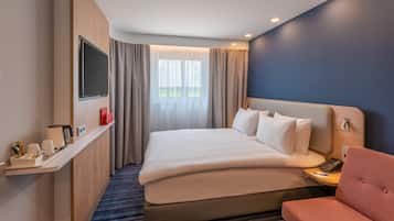 Una habitación de hotel con una cama grande, un televisor montado en la pared, una mesita de noche con un teléfono y una lámpara, y una ventana con cortinas.