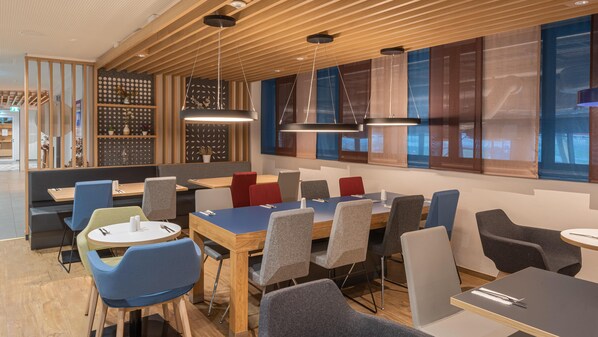A modern restaurant with wooden ceiling panels, large windows with blue curtains, and a variety of seating options including booths and tables. - Holiday Inn Express Hamburg South A7 As42 (Egestorf)