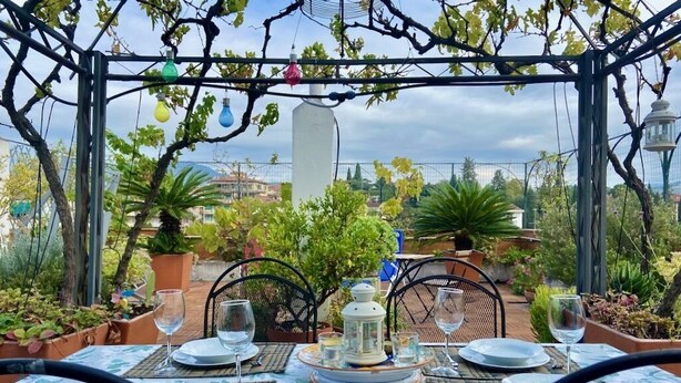 Un espace de restauration en plein air avec une table dressée pour deux personnes, entourée de chaises, sous une pergola ornée de guirlandes lumineuses.