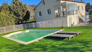 A swimming pool with a grassy lawn and lounge chairs, surrounded by a fence and a building with a balcony. - VILLA 14 Pers. Private pool, pétanque, terrace. 1km from Vallon center (Vallon-Pont-d'Arc)