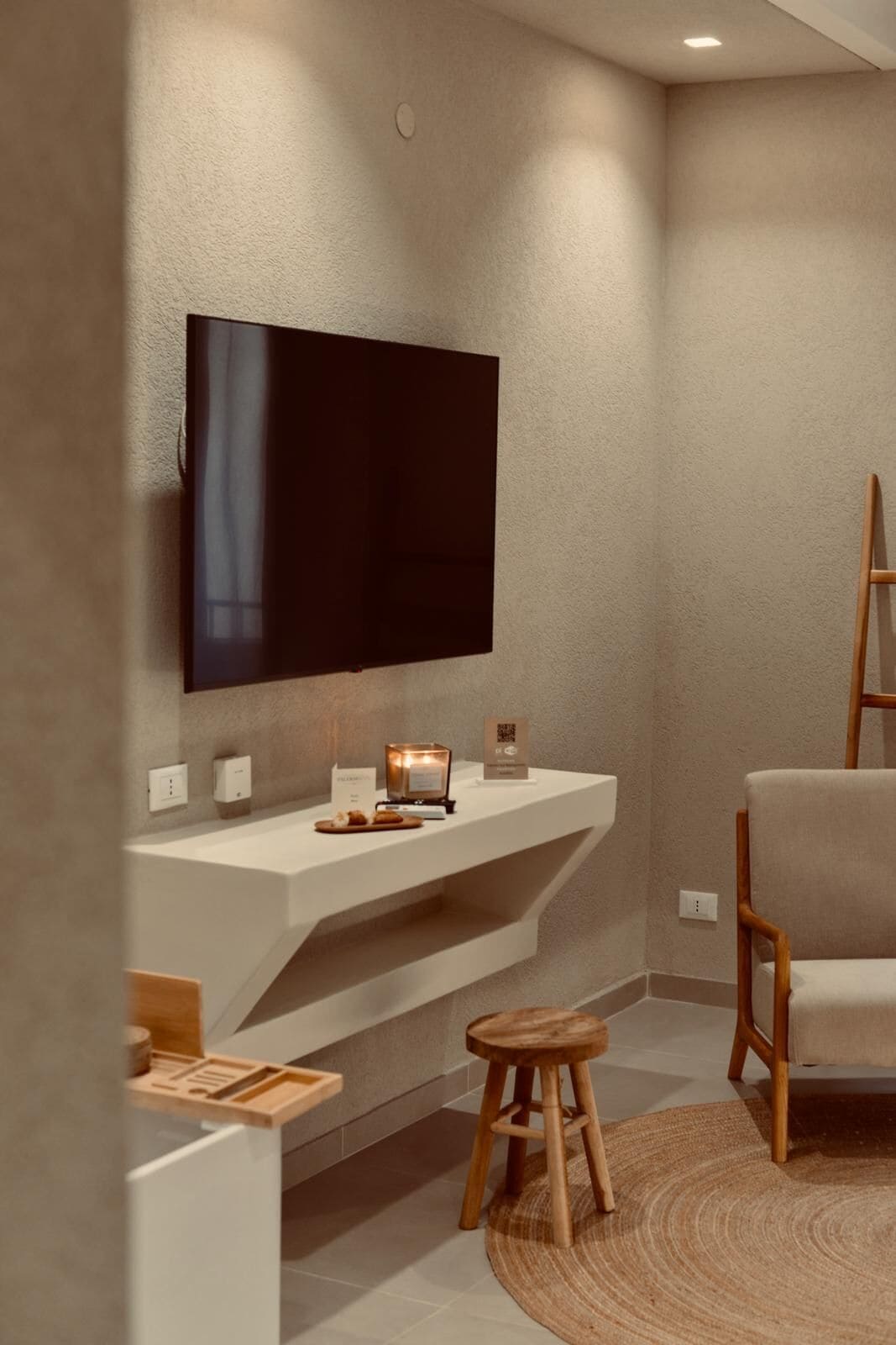 A modern living room with a flat-screen TV mounted on a textured wall, a wooden chair, a small stool, and a white console table with a candle and a QR code sign.