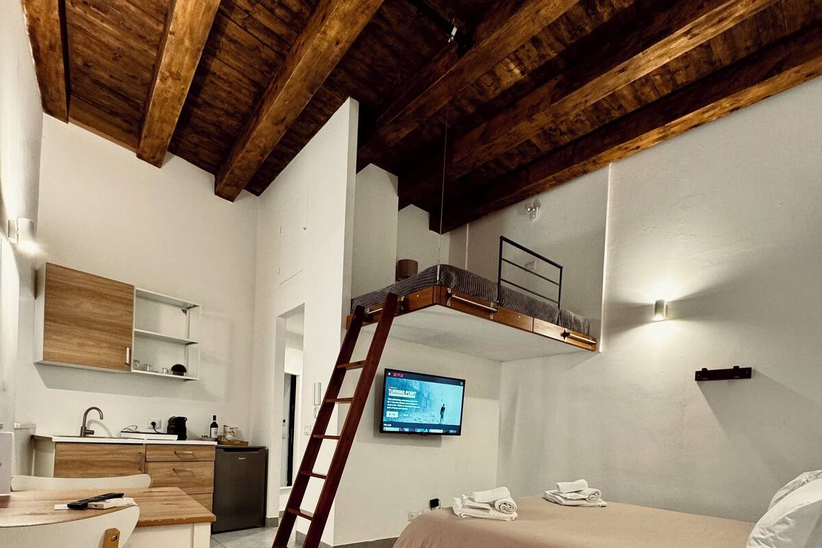 A compact room with a bunk bed, a desk with a chair, a small kitchenette, and a television mounted on the wall.
