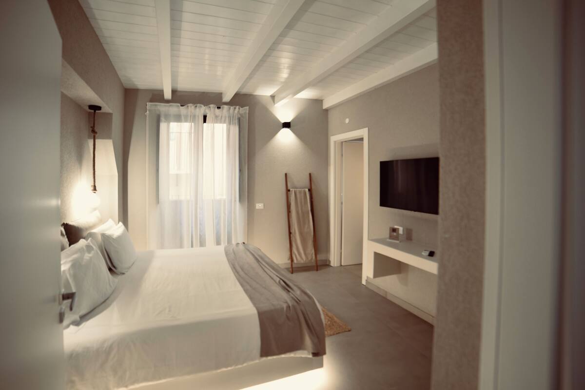 A modern bedroom with a large bed, a TV mounted on the wall, and a window with curtains.