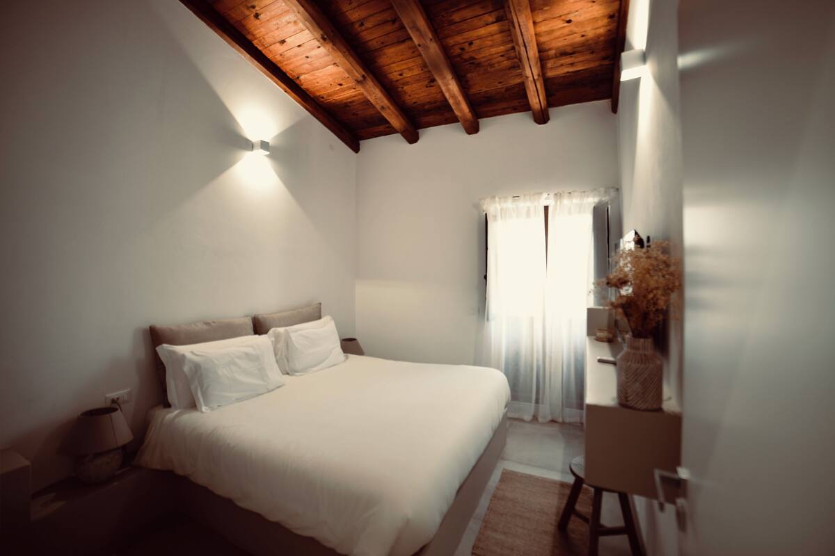 A bedroom with a wooden ceiling, a bed with white bedding, a nightstand, a lamp, and a window with sheer curtains.