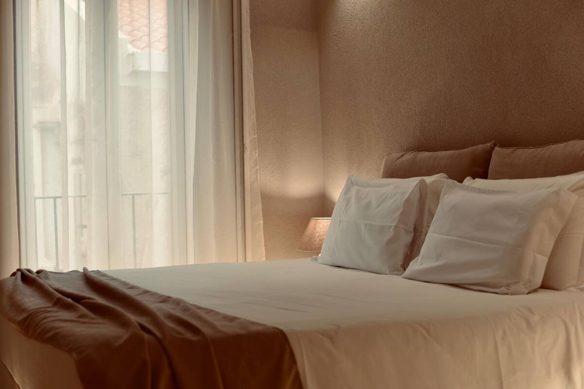 A neatly made bed with white linens and a brown throw blanket, a bedside lamp, and a view of a building through sheer curtains.