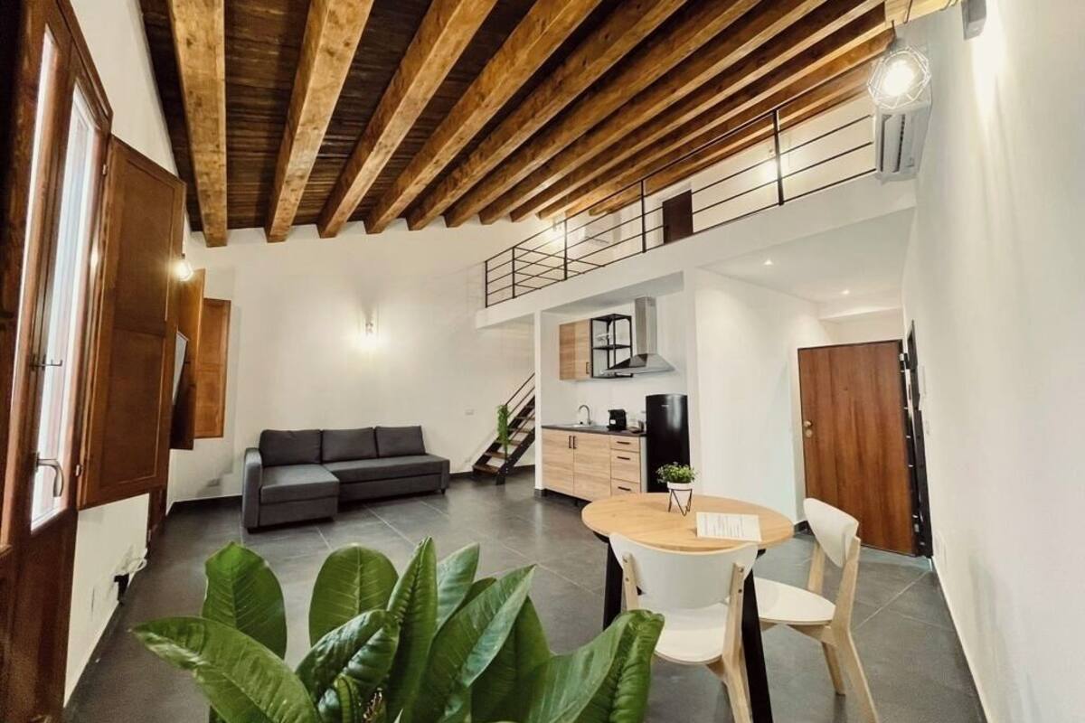 A modern interior with a wooden ceiling, a dining area with a round table and chairs, a grey sofa, and a kitchen in the background.