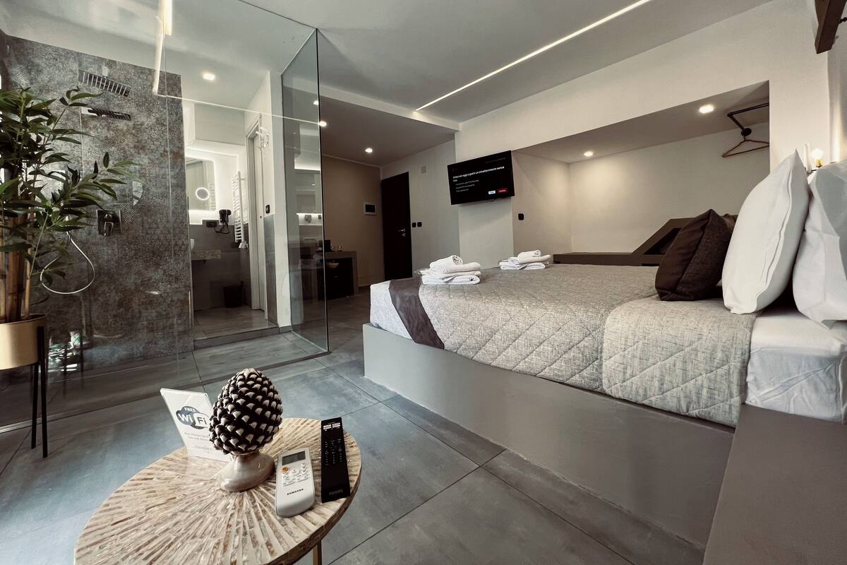 A modern hotel room with a large bed, a TV mounted on the wall, and a glass partition separating the bedroom from the bathroom.