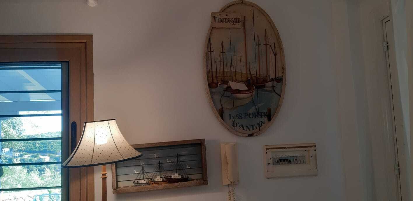 A room with a wooden door, a wall-mounted lamp, a framed maritime painting, and a small electrical panel.
