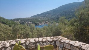 A stone wall with a cactus in the foreground, a view of a coastal village with buildings and a body of water, and a hill in the background. - Eumaeos Spitakia (Σύβοτα Λευκάδας)