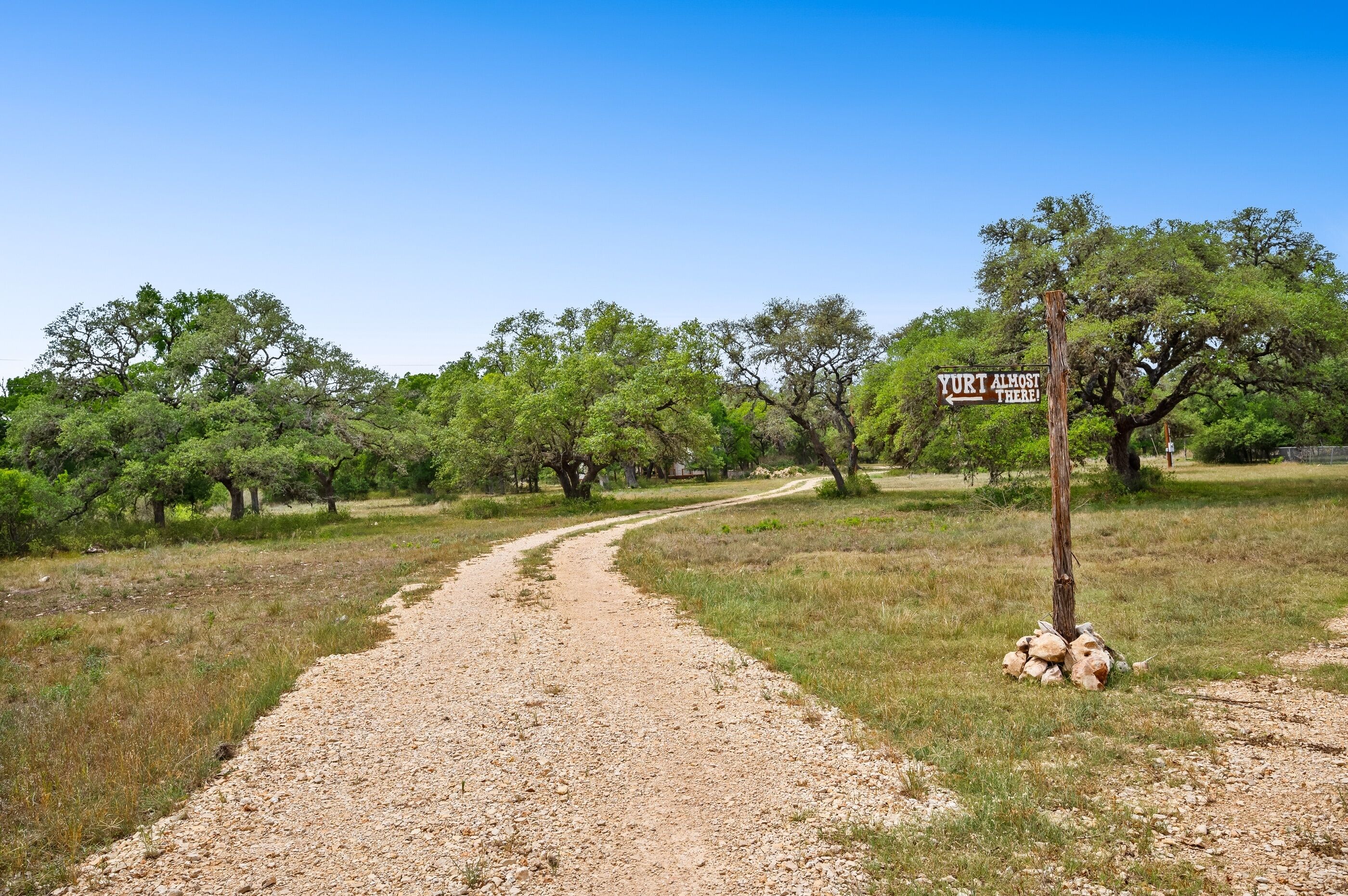 Un sendero de grava que serpentea por un área boscosa, con un letrero que dice '¡Yurta casi a la vista!'.