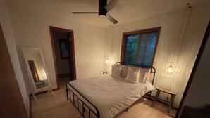 A bedroom with a bed, wooden floor, ceiling fan, bedside lamps, and a window with a view of trees. - Lakes and Beaches with Resort Amenities at The Hideout (Hamlin)
