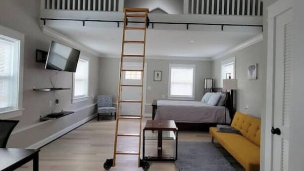 A loft-style bedroom with a loft bed, a desk, a chair, a TV mounted on the wall, and a window.