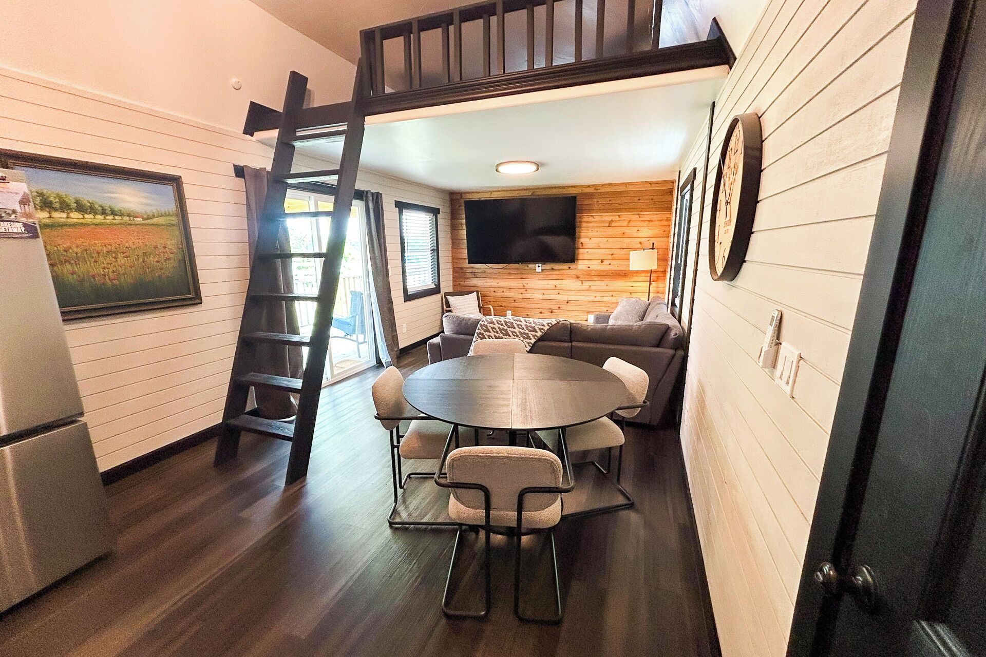 A compact living space with a dining area, a sofa, a TV mounted on the wall, and a ladder leading to an upper level.