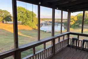 Una terraza de madera con vista a un lago y barcos, un área con césped y árboles.