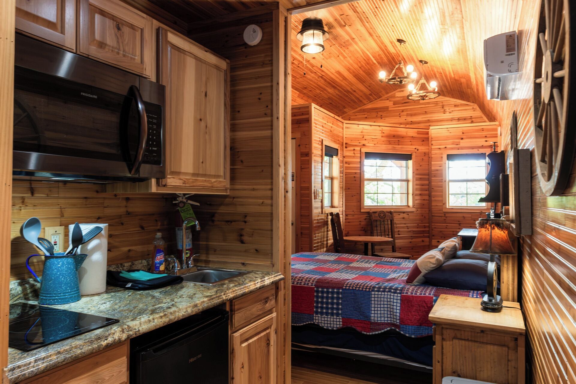 L’intérieur chaleureux d’un chalet avec des murs en bois, un coin cuisine équipé d’un four à micro-ondes et d’une cuisinière, une table à manger avec des chaises et un lit recouvert d’une courtepointe.