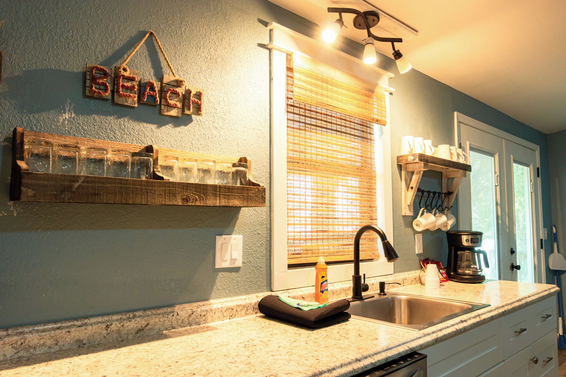 A kitchen with a wooden shelf holding glasses, a sink with a faucet, and a window with blinds.