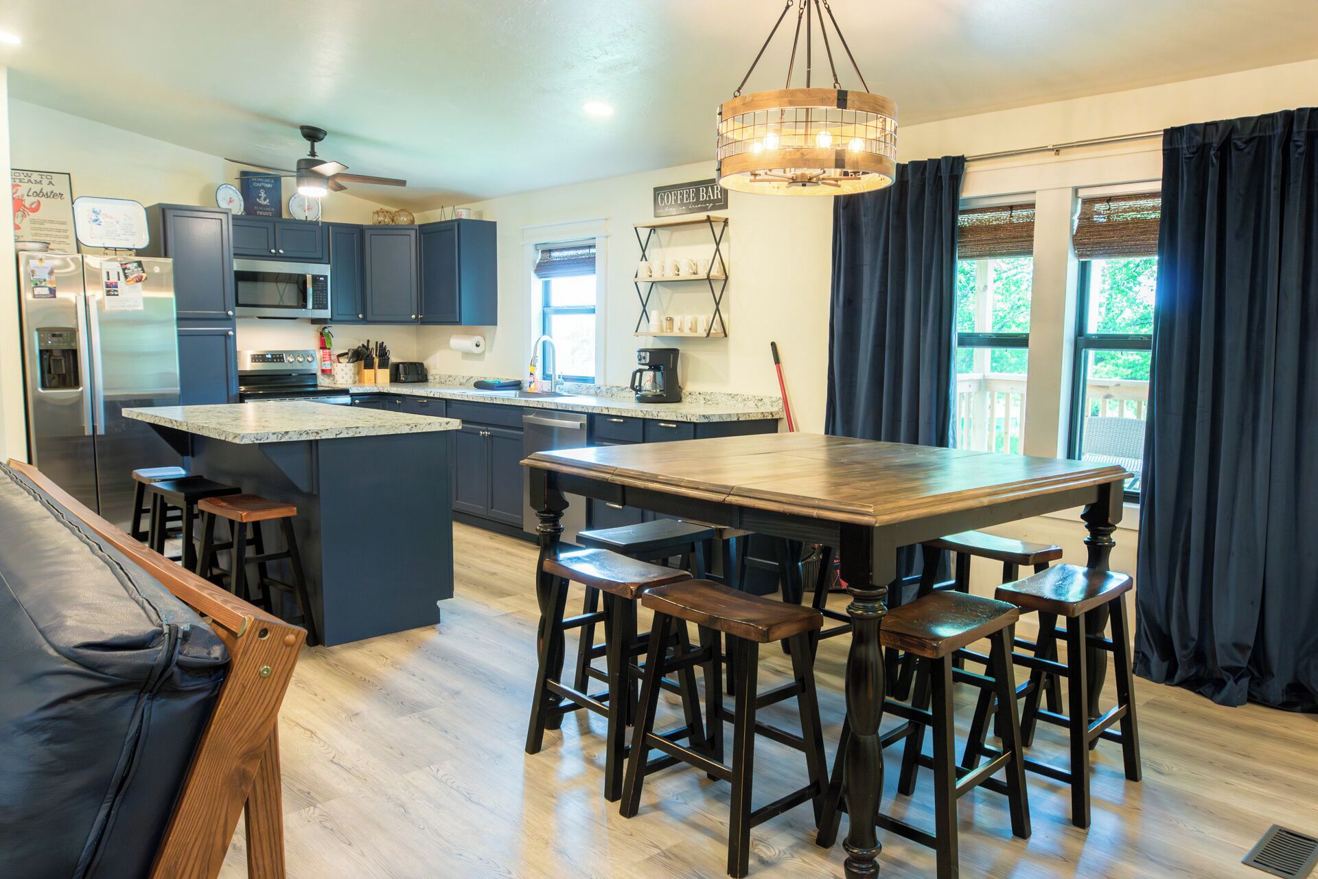A modern kitchen with dark blue cabinets, a large island with stools, a coffee bar, and a view of greenery outside.