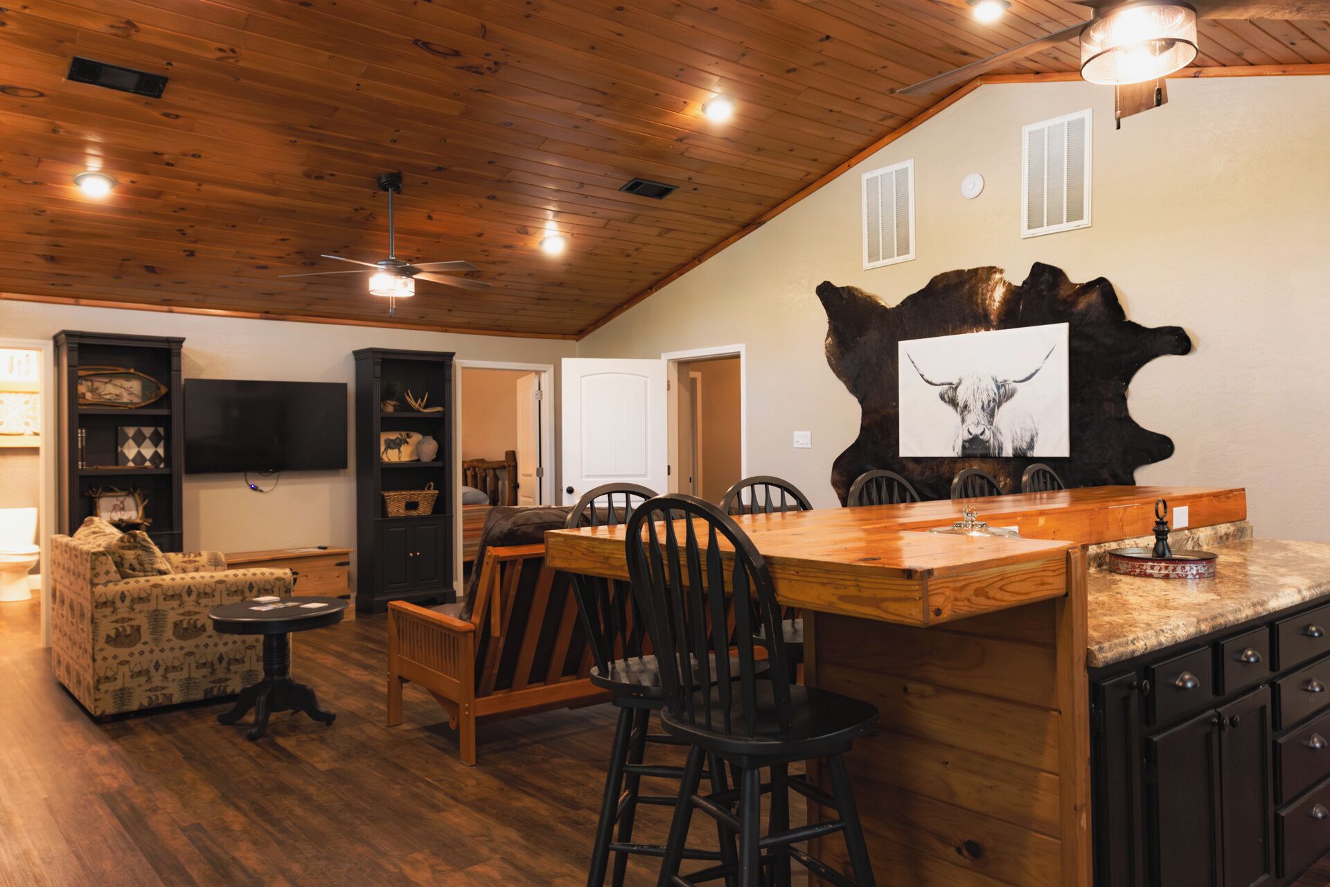 A spacious living area with a wooden ceiling, a dining table with chairs, a bar area, and a television mounted on the wall.