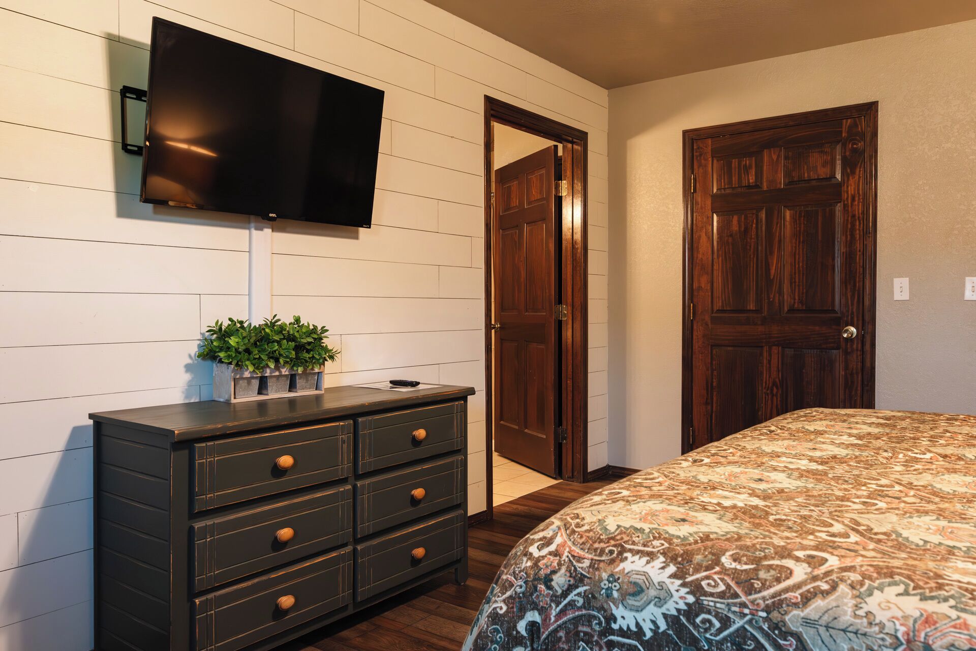Un dormitorio con cama, cómoda, televisor montado en la pared y dos puertas de madera.