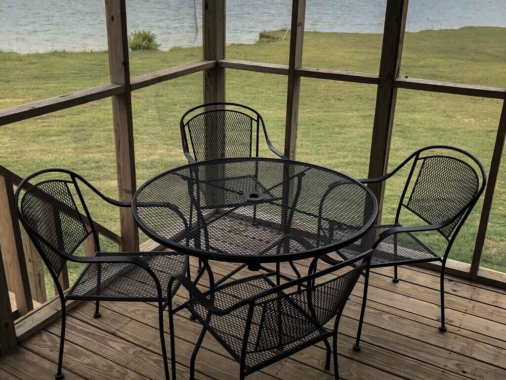 A covered outdoor area with a round metal table and four chairs, overlooking a grassy area and a body of water.