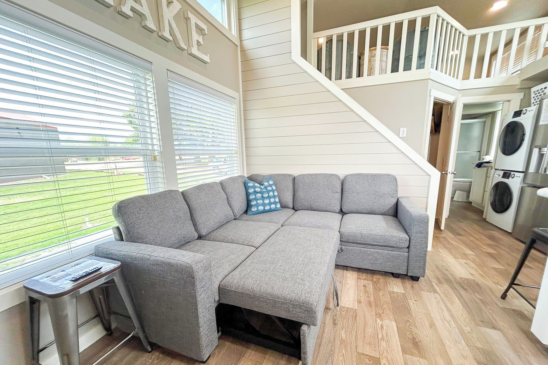 Un canapé d’angle gris avec un coussin bleu, une petite table avec une télécommande et un escalier menant à une buanderie.