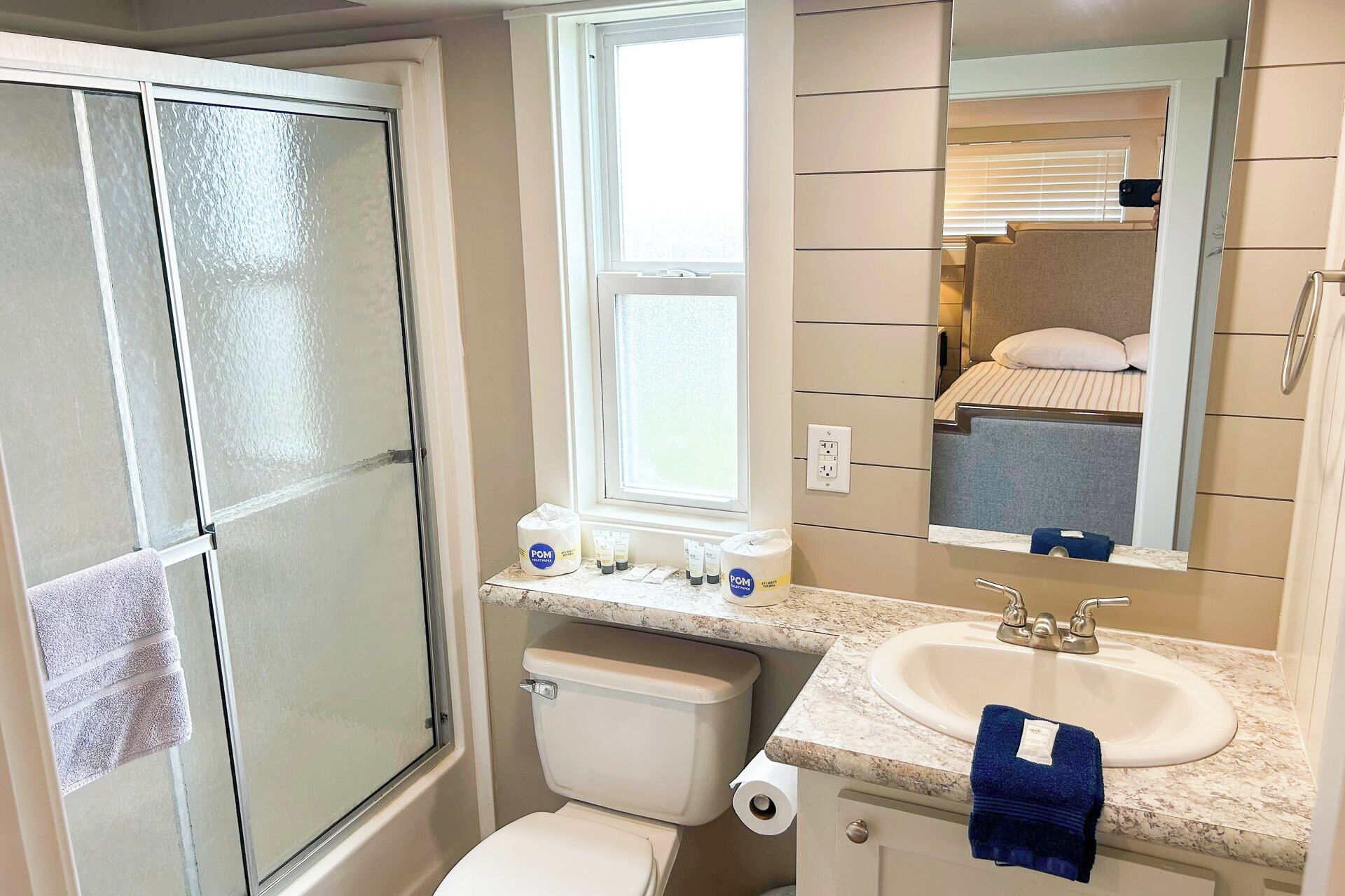 Une salle de bain équipée d’une douche, de toilettes, d’un lavabo et d’un miroir dans lequel on peut voir un lit.