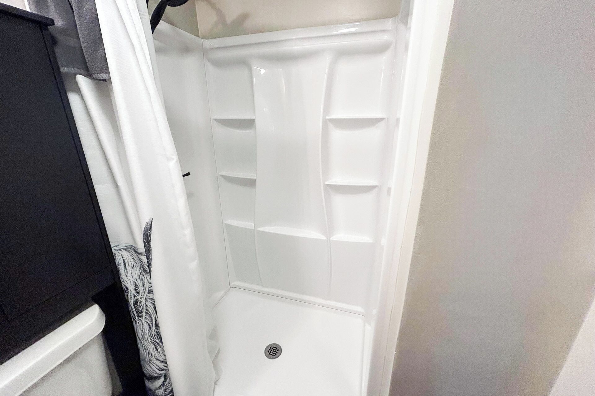 A white shower with a drain in a corner, a white wall, and a black object partially visible on the left.