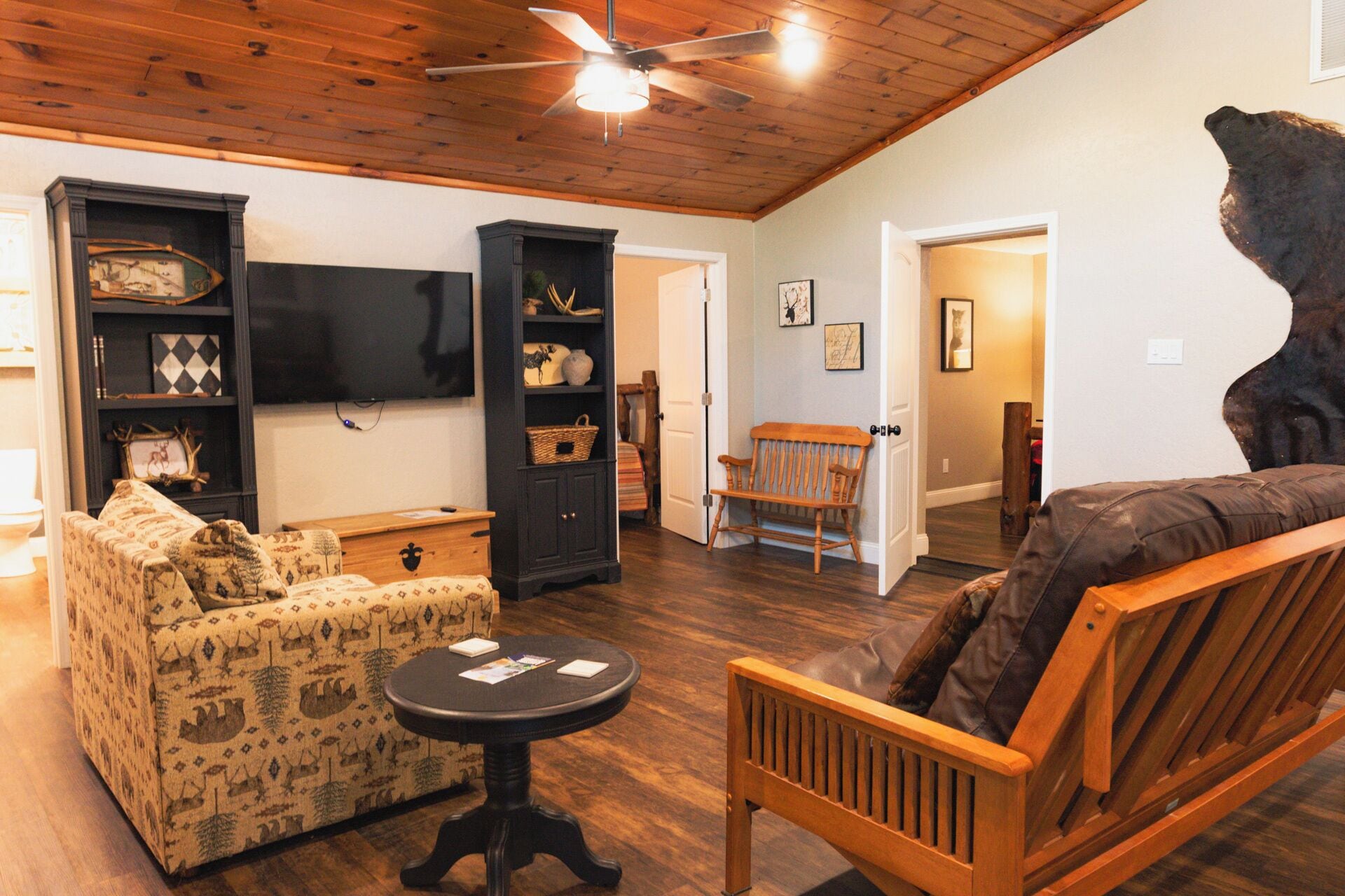 A cozy living room with a wooden ceiling, a television mounted on the wall, a sofa, a rocking chair, and a small table with magazines.