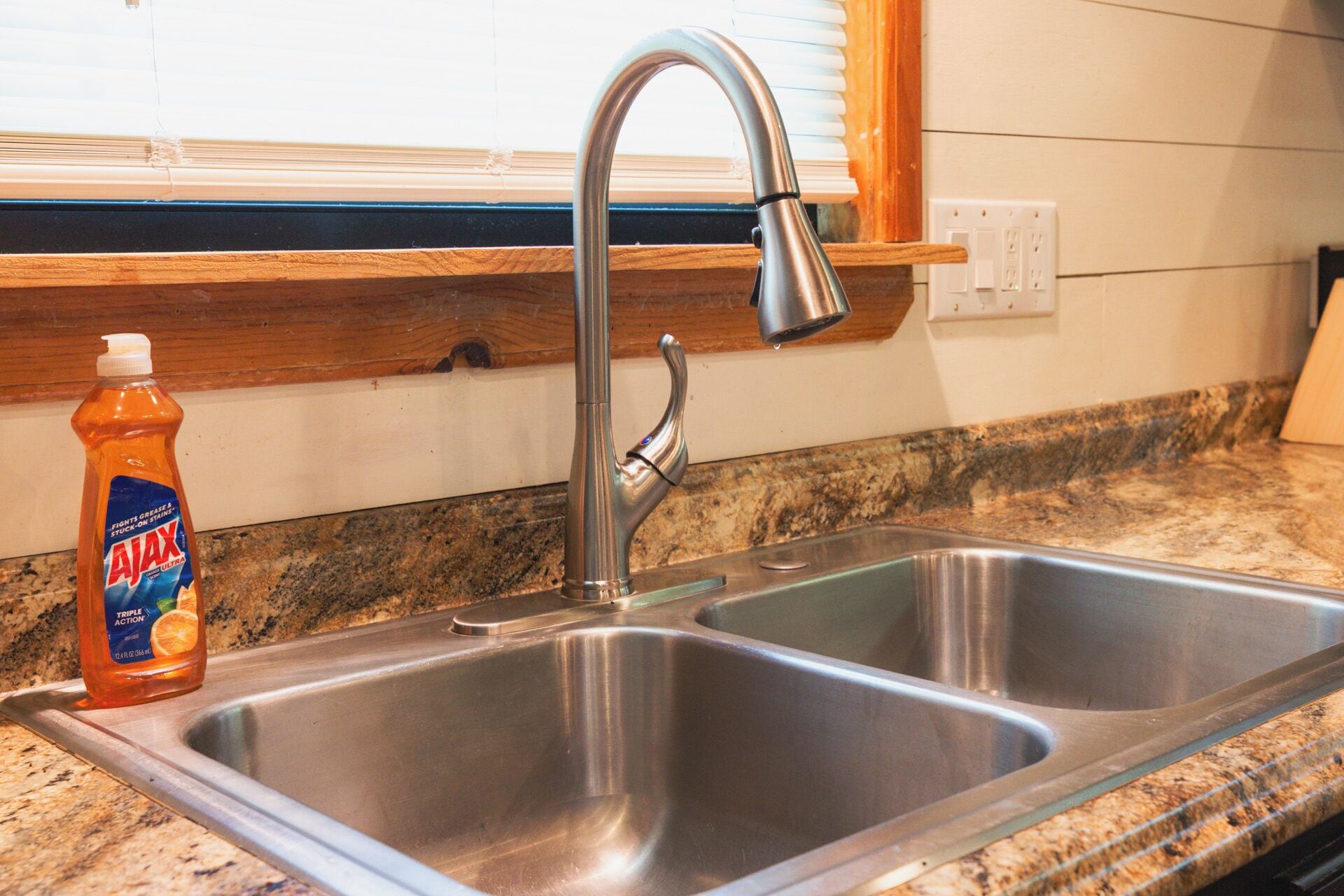 A kitchen with a stainless steel sink, a bottle of Ajax dish soap, and a window with blinds.