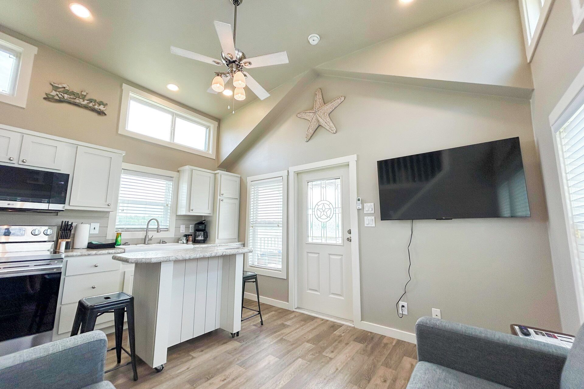 A modern kitchen with a central island, a dining area with a grey sofa, and a flat-screen TV mounted on the wall.
