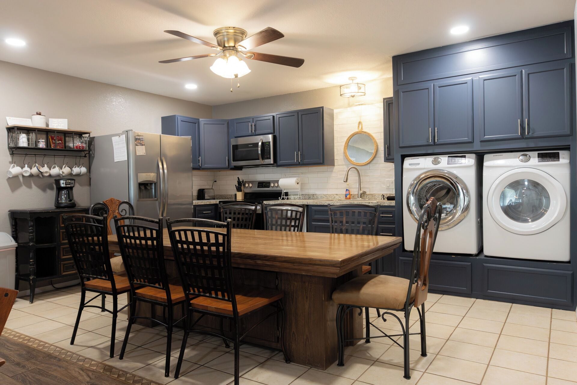 A modern kitchen with dark blue cabinets, a central island with chairs, a ceiling fan, a microwave, a refrigerator, and a washing machine.