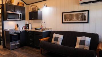 A compact kitchen with a microwave, stove, and sink. There is a framed picture on the wall and a dark sofa with cushions.