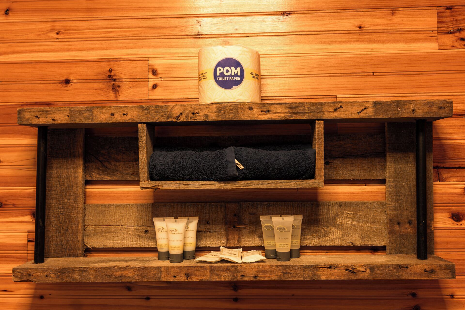 A wooden shelf with a roll of POM toilet paper, a black towel, and three tubes of Nove Fresh Screen shampoo.