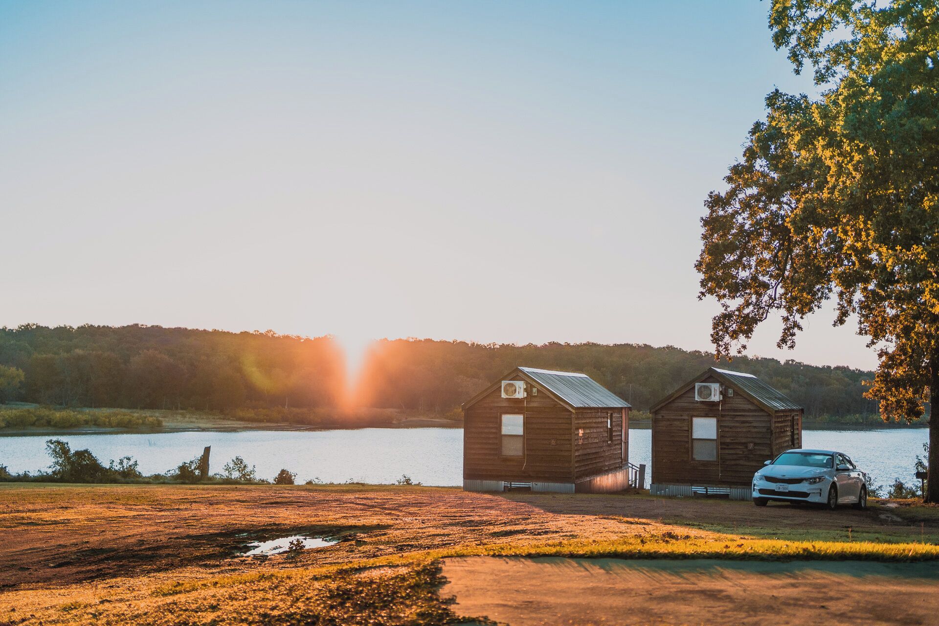 Une cabane rustique avec une voiture garée devant, située au bord d’un lac, avec un coucher de soleil en arrière-plan.