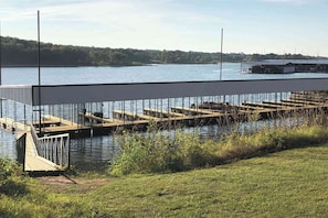 Un muelle que se adentra en un lago, con un área verde al frente y un edificio en la orilla opuesta.