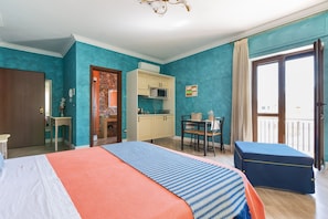 A hotel room with a bed, a desk, a kitchenette, and a balcony.