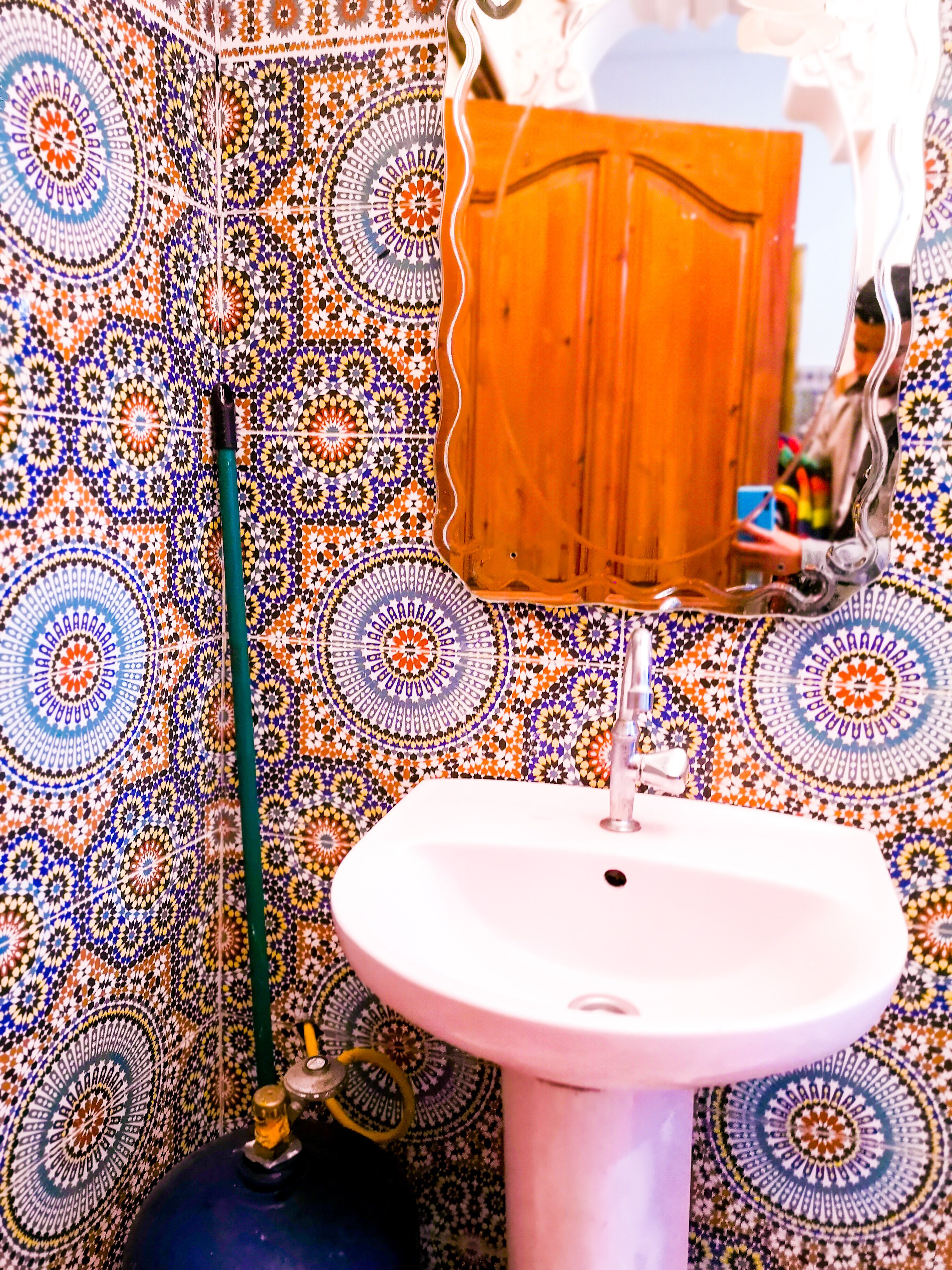 A bathroom with a patterned tile wall, a sink, and a mirror reflecting a wooden door.