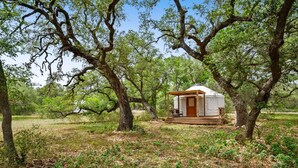 A rustic cabin with a wooden deck and a covered porch, surrounded by dense trees and a grassy area. - Ot 3515e Texas Yurt Haus: 5 Yurts! 5 Bedroom Cabin by RedAwning (New Braunfels)