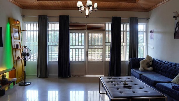 Un salon spacieux avec un plafond en bois, une grande fenêtre avec des rideaux, un canapé bleu, un pouf capitonné et un ventilateur.