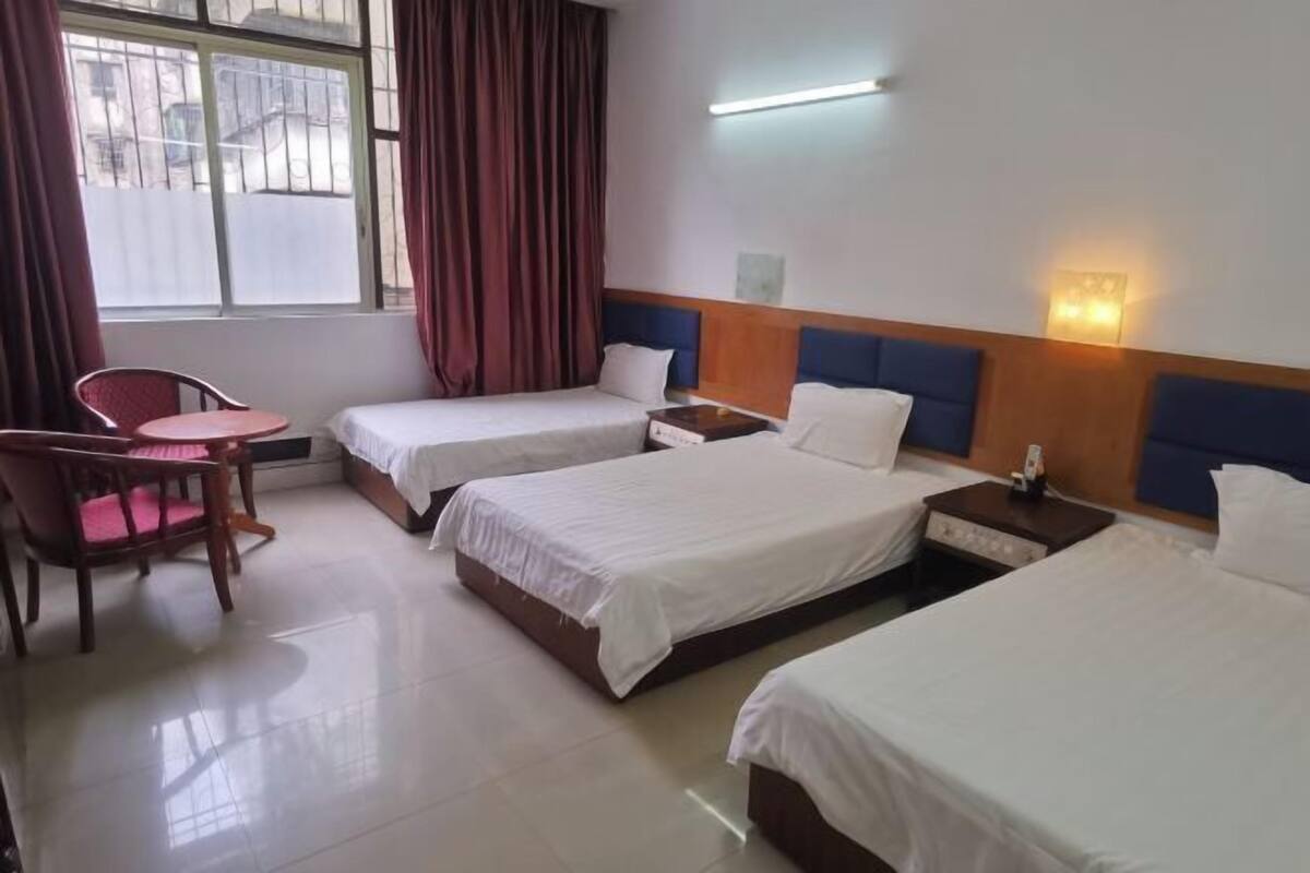 A hotel room with three single beds, a small table, a chair, and a window with curtains.