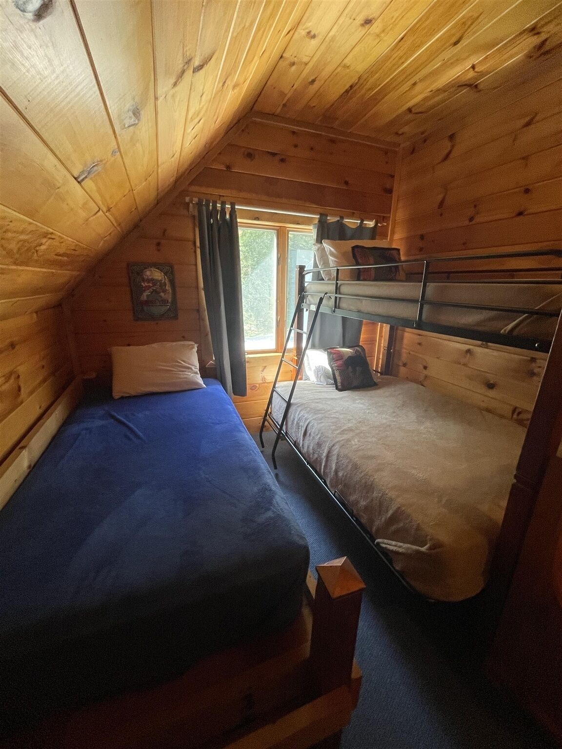 A small, cozy room with wooden walls and a sloped ceiling. It features two bunk beds with blue and beige bedding, a small window with curtains, and a framed picture on the wall.