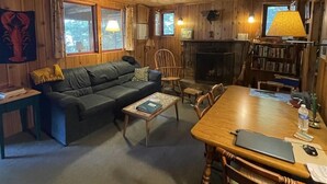 A cozy cabin interior with a wooden ceiling, a fireplace, a sofa, a dining table, and a bookshelf. - Greens Limit 6 2 Bedroom Cottage by RedAwning (Sebago)