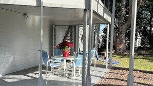 En overdækket terrasse med et bord og stole, en potteplante samt udsigt til en boligbygning og et træ.