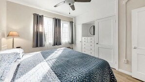 Une chambre à coucher avec un lit, un ventilateur de plafond, une fenêtre avec des rideaux, une commode et une porte.