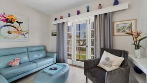 A modern living room with a blue sofa, a grey armchair, and a coffee table. There is a large window with curtains, a painting of a bicycle, and a shelf with decorative items. - Charming Sweet Daisy overlooking stunning new pool in awesome Southside Village! (Sarasota)
