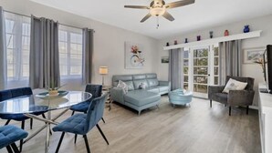 A modern living room with a glass dining table, blue chairs, a grey sofa, and a ceiling fan. - Charming Sweet Daisy overlooking stunning new pool in awesome Southside Village! (Sarasota)