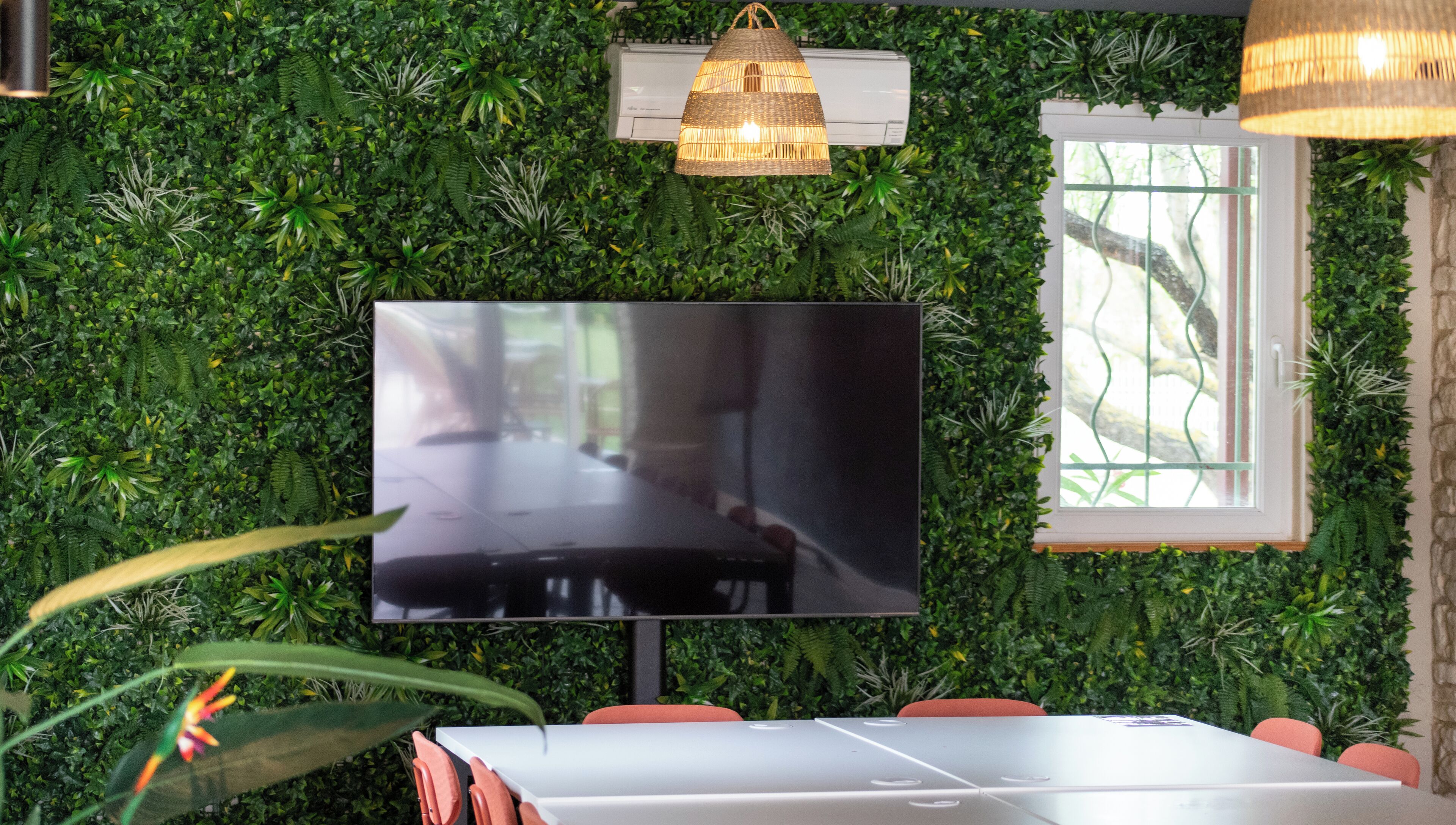 Quarto com uma grande televisão de tela plana fixada em uma parede coberta de folhagem verde, janela com vista para árvores e luminária pendente.