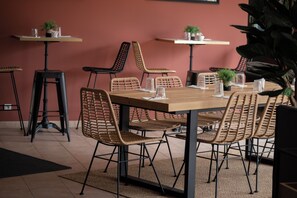 Un comedor con una mesa de madera larga, sillas de mimbre y taburetes negros. Hay pequeñas mesas con plantas en macetas y cristalería en las paredes.
