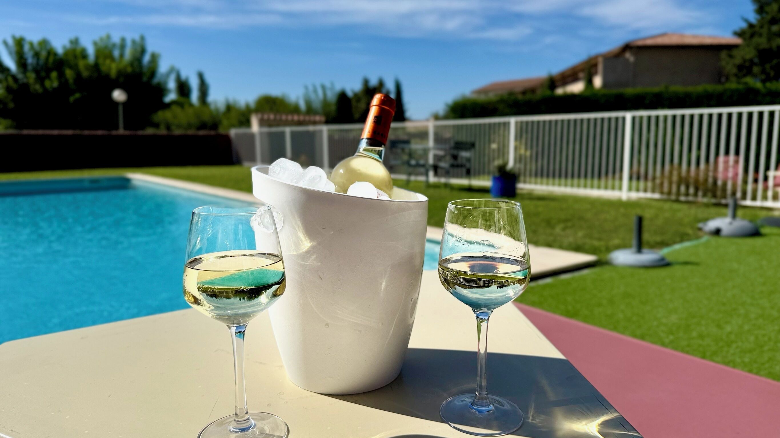 Escena junto a la piscina con un cubo blanco que contiene una botella y hielo, y dos copas de vino blanco sobre una mesa.