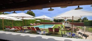 Un área de piscina exterior con múltiples sillas de descanso y sombrillas, una zona de barbacoa y un césped bien cuidado.